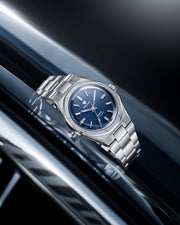 Nivada Grenchen F77 Blue MK2 automatic watch with textured blue tapisserie dial SST displayed on reflective metallic surface