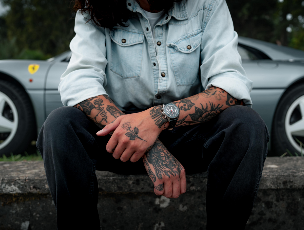 A tattooed man wearing the watch rests his forearms on his knees beside a parked sports car.