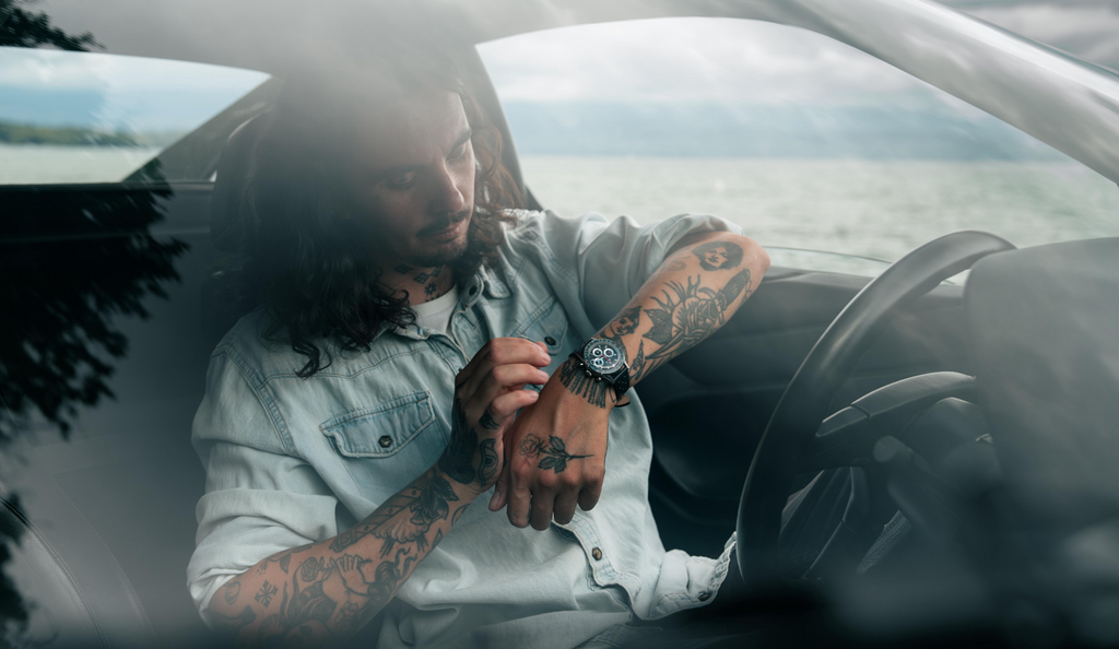 A man adjusts the watch on his wrist while seated in a convertible beside a lake.