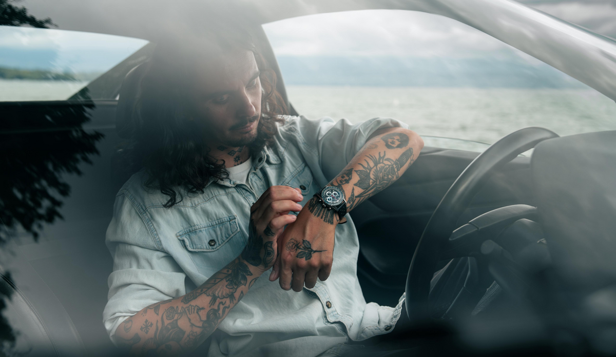 A man adjusts the watch on his wrist while seated in a convertible beside a lake.
