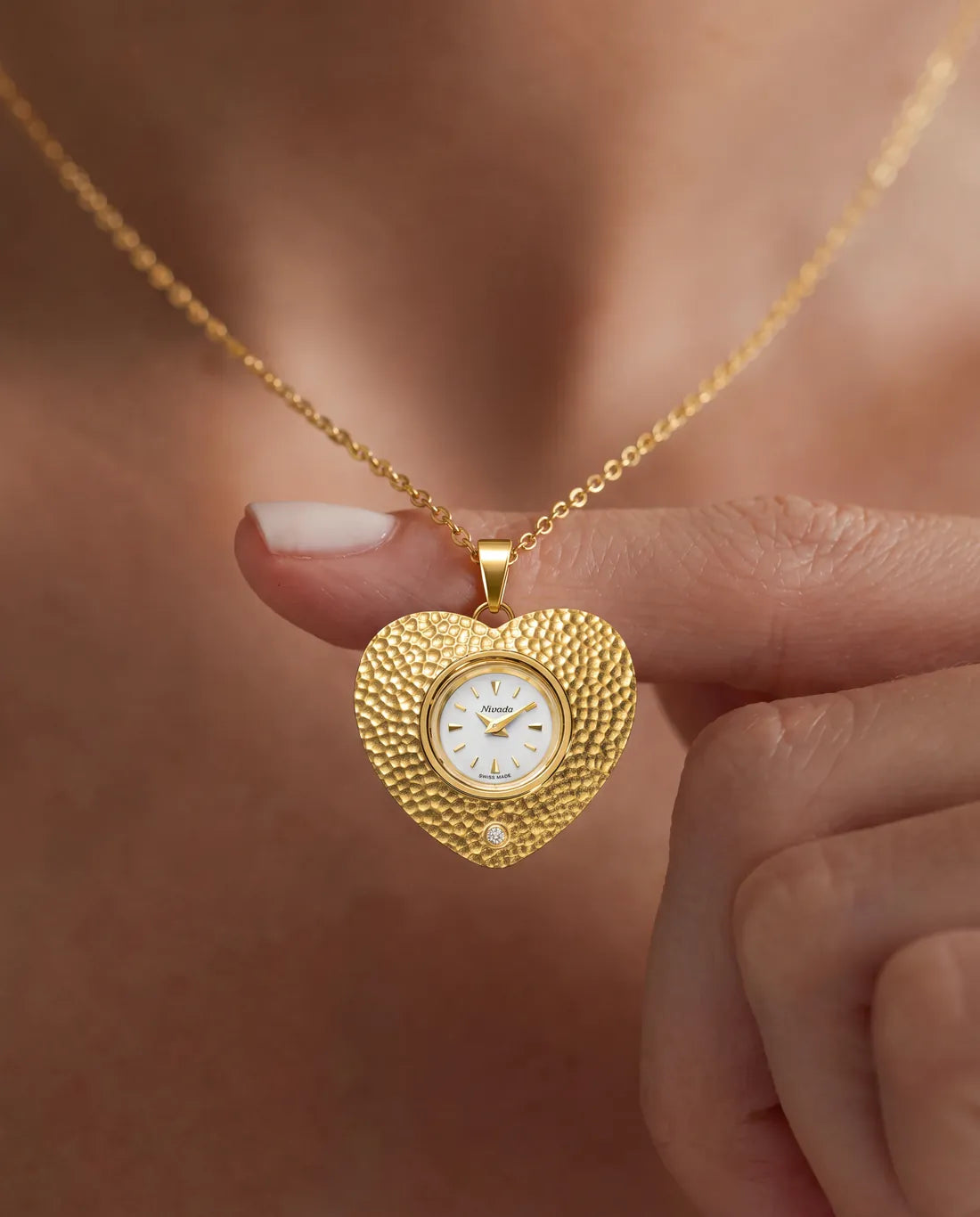 The Colorama heart pendant, hammered gold with a diamond set below the dial, held on a fingertip