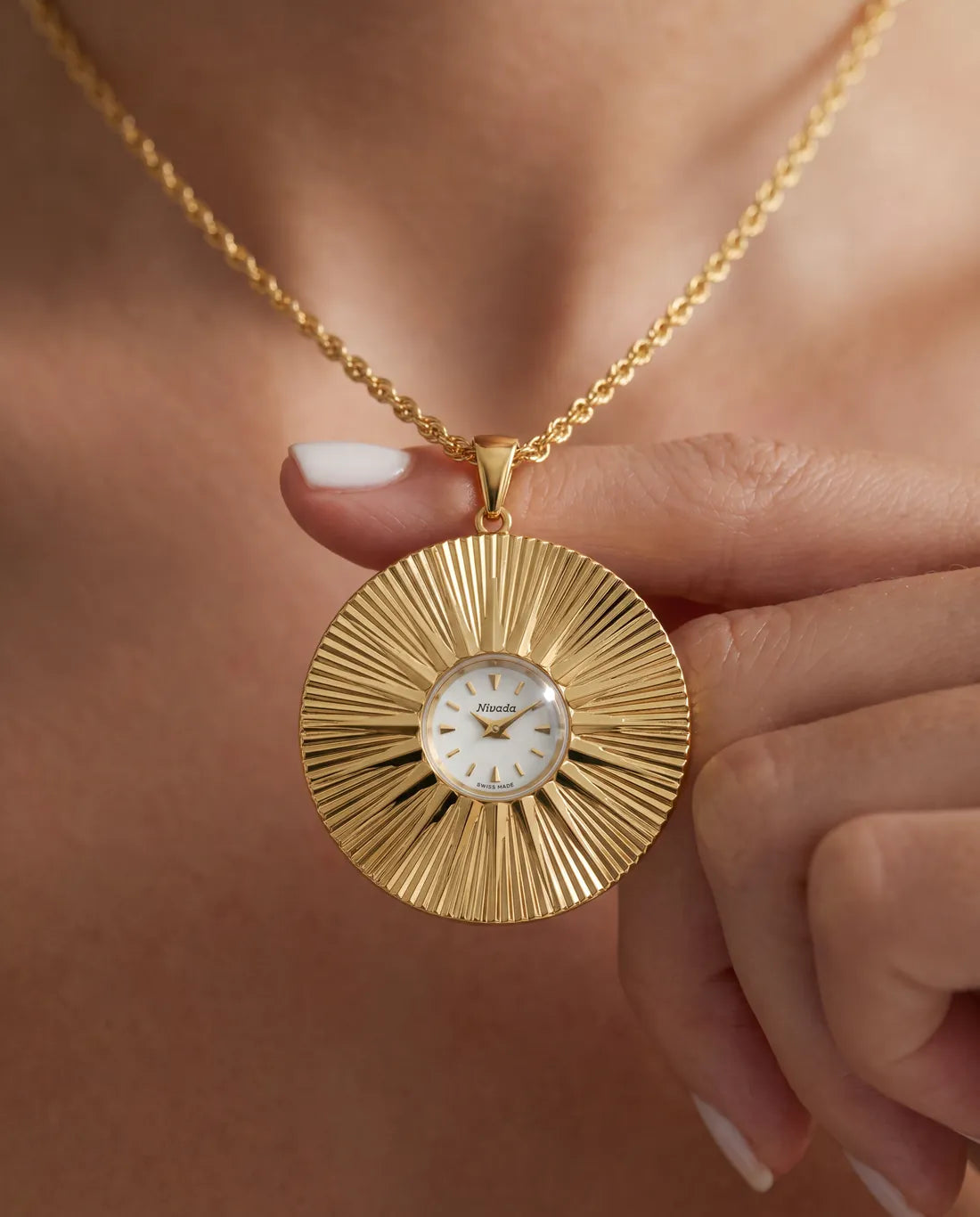 The Colorama gold pendant, a sunburst of fluted gold around a small white Nivada dial, held on a fingertip